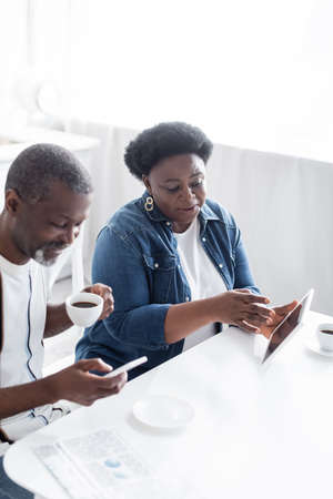 african american woman holding digital tablet near cheerful husband with cup of coffee  using smartphoneの写真素材
