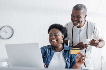 senior african american woman holding credit card near laptop and happy husband with cupの写真素材