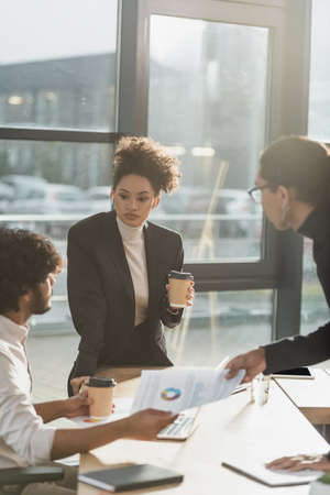 African american businesswoman holding coffee to go near blurred colleagues with paper in office