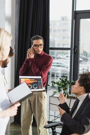 African american businessman talking on smartphone near interracial colleagues with paper and notebook in officeの写真素材