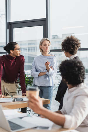 Young businesswoman talking to multiethnic colleagues near papers and laptop in office