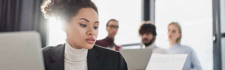 Young african american businesswoman looking at paper near blurred laptop and colleagues in office, bannerの写真素材