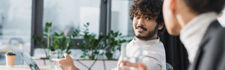 Indian businessman pointing at laptop near blurred african american colleague in office, banner