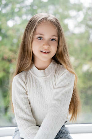 positive girl with long hair smiling at camera near blurred window