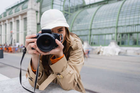 young photographer in trench coat and baseball cap taking photo on digital camera