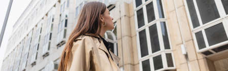 low angle view of young woman in trendy trench coat looking at building, banner