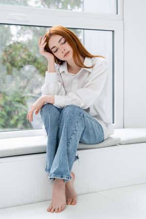 Barefoot woman in shirt and jeans sitting on windowsill at homeの写真素材