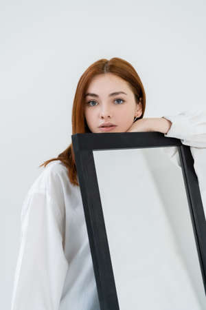 Redhead woman in shirt looking at camera near mirror isolated on white