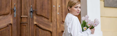 happy young woman with bouquet of fresh tulips near blurred wooden doors, bannerの写真素材