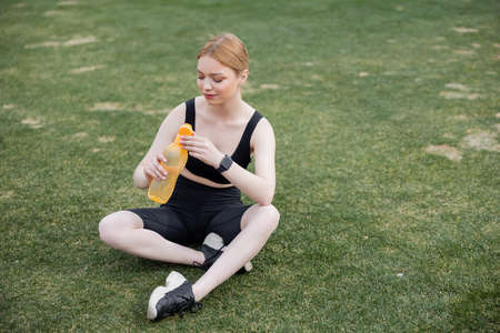 young sportswoman sitting on stadium and opening sports bottleの写真素材