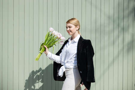 young woman in white shirt and black blazer walking with tulips near grey wallの写真素材