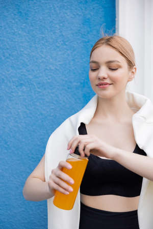 young and happy sportswoman opening bottle with orange juice near blue wall