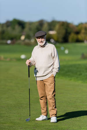senior man in flat cap standing with hand in pocket and golf club on green fieldの写真素材