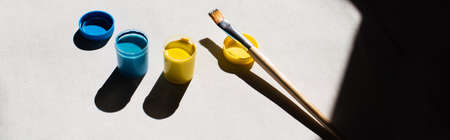 high angle view of blue and yellow paint in jars near paintbrush on white, bannerの写真素材