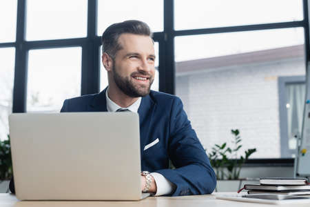 cheerful economist looking away while working near laptop and notebooks