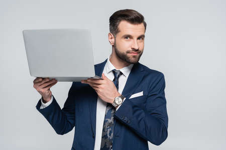 positive businessman looking at camera while holding laptop isolated on greyの写真素材