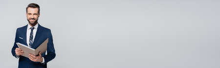 smiling businessman in blue blazer holding folder and looking at camera isolated on grey, bannerの写真素材