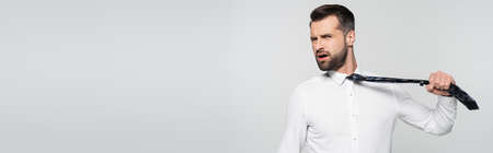 irritated businessman pulling tie while looking at camera isolated on grey, bannerの写真素材