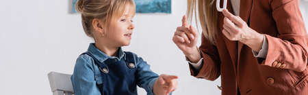 Smiling girl pointing with finger near speech therapist with letter in consulting room, banner