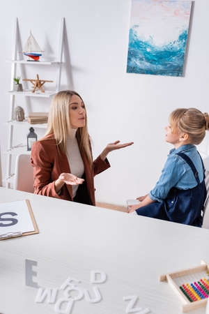 Speech therapist talking with kid near letters and clipboard on table