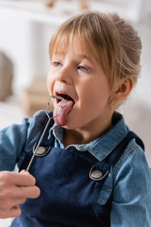 Speech therapist holding logopedic probe near kid sticking out tongue in classroom
