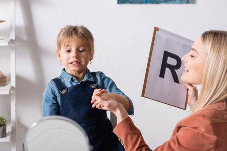 Smiling speech therapist holding clipboard and hand of child near mirror in consulting roomの写真素材