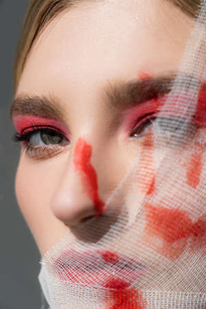 Close up view of woman with red paint on medical bandage on face looking at camera isolated on grey