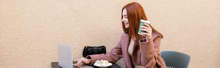 happy freelancer holding coffee to go and using laptop near plate with macarons, banner