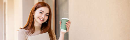 happy young woman with red hair holding laptop and paper cup near building, banner