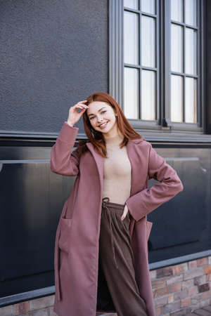 cheerful redhead woman in stylish coat standing with hand on hip near modern buildingの写真素材