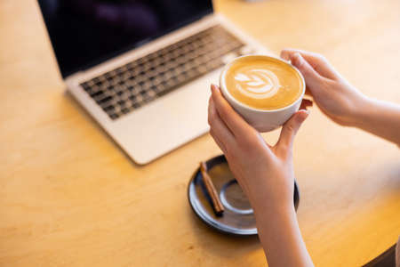 cropped view of young freelancer holding cup of cappuccino near laptop in cafeの写真素材