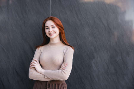 happy redhead woman in beige turtleneck standing with crossed arms near grey marble wallの写真素材