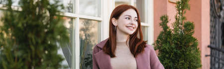 young pleased woman with red hair smiling near building, bannerの写真素材