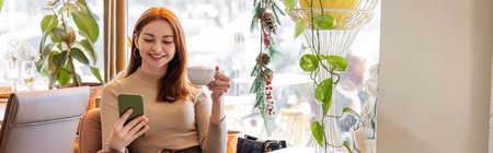 happy young woman with red hair using smartphone and holding cup of coffee, banner