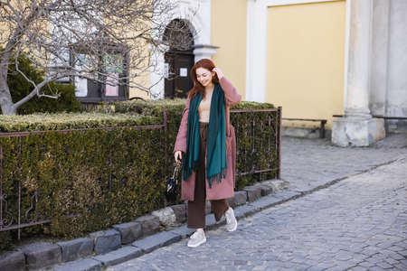 full length of positive young woman in coat and scarf holding handbag and walking on street
