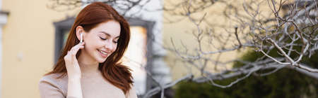 happy and redhead woman adjusting wireless earphone outside, banner
