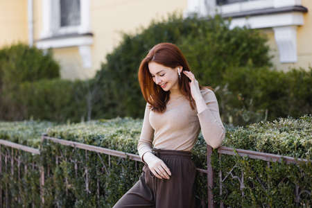 young pleased woman in wireless earphone adjusting red hair outside