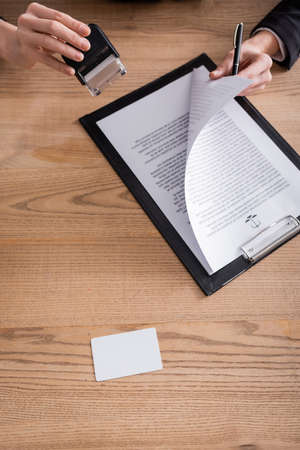 top view of contract and blank business card near cropped notary with stamper and pen