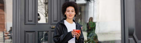pleased african american woman in jacket holding cup of coffee on street, banner