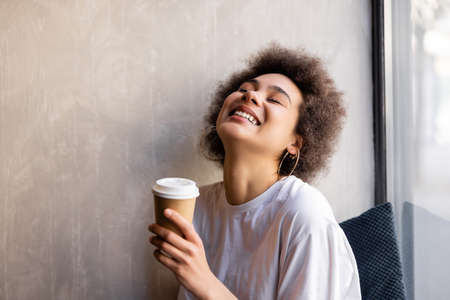 pleased african american woman in white t-shirt holding paper cup