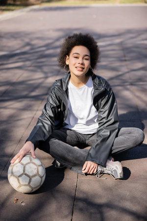 full length of smiling african american woman in jacket sitting with crossed legs and ball on asphaltの写真素材