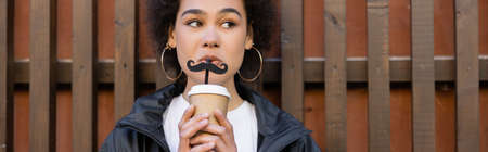 curly african american woman drinking takeaway beverage from straw with moustache near fence, bannerの写真素材