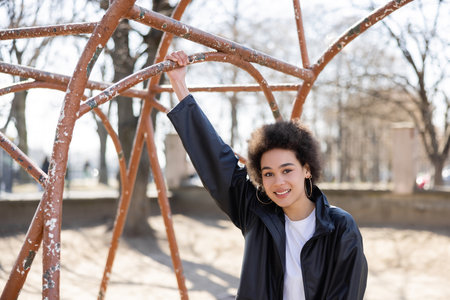 joyful african american woman in jacket smiling at rusty outdoor gym