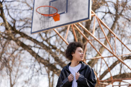 low angle view of happy african american woman in jacket standing on basketball outdoor court
