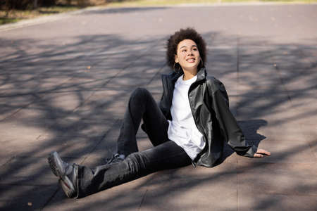 full length of pleased african american woman in hoop earrings and jacket sitting on asphalt