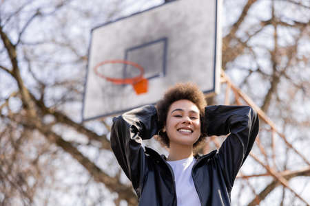 low angle view of cheerful african american woman in jacket standing on basketball outdoor courtの写真素材