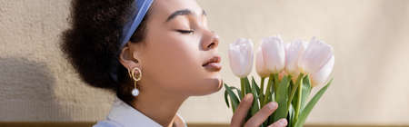 curly african american woman in blue headband smelling tulips near wall, bannerの写真素材