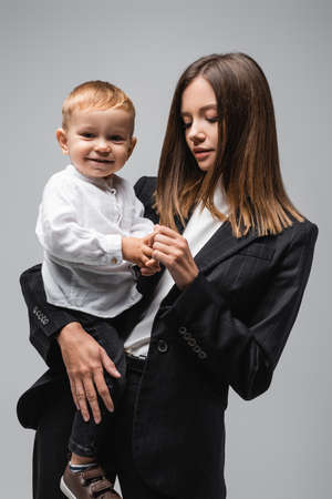 young woman in black suit holding hands of happy son isolated on greyの写真素材