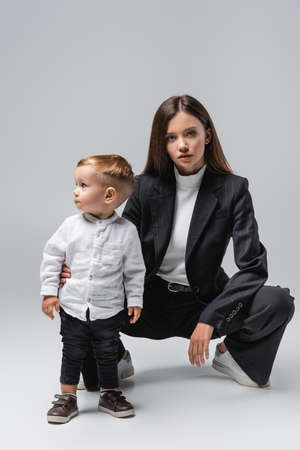 young woman in black suit looking at camera while posing near toddler son on greyの写真素材