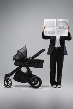 woman in black suit obscuring face with newspaper near baby carriage on greyの写真素材